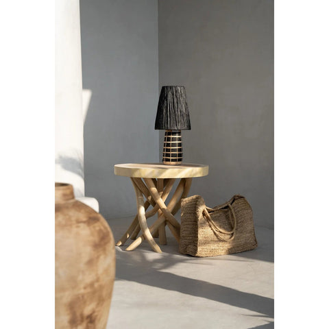 A round wooden side table with curved legs features The Naxos Table Lamp - Black Natural, while a woven rattan bag rests on the floor nearby, all against a minimalist, sunlit wall.