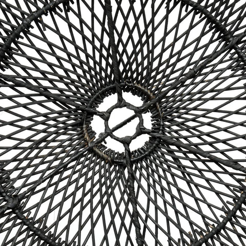 An overhead close-up of The Chapeau Pendant - Black - L showcases its circular, woven wire design with a geometric pattern, resembling a rattan lamp shade. Black wires radiate from the center against a white background.