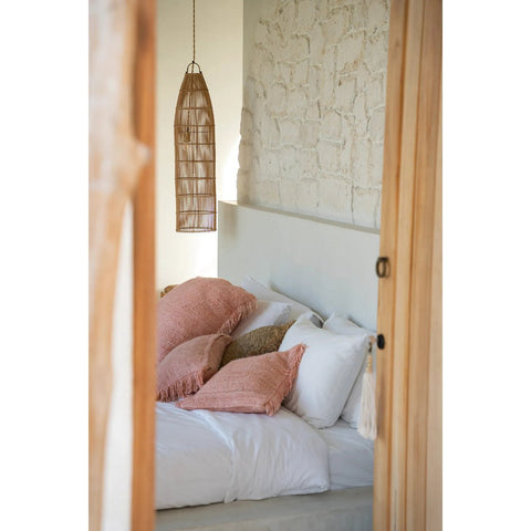 A cozy bedroom with a white bed, pink and beige textured pillows, a rustic white brick accent wall, light wooden door frames, and The Fish Trap Pendant - Natural - L as a woven boho rattan lamp.