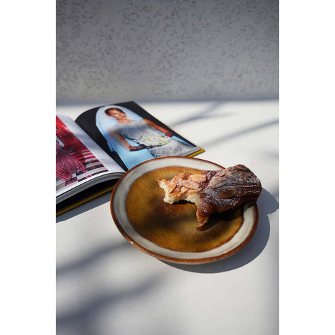 A partially eaten pastry sits on a brown ceramic plate with a crackle glaze finish beside an open magazine featuring a model in a white dress, all on a sunlit white surface with soft shadows.