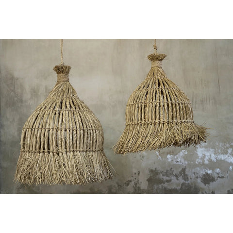The Guagua Pendant - Natural - L: A pair of hanging woven straw lampshades with fringes, set against a textured gray concrete wall. These handmade grass pendants offer rustic charm and natural fiber details for an organic look.