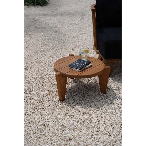 The Seseh Coffee Table - S - Outdoor sits on gravel, holding a glass of lemon water and a closed book. A black cushioned chair is partially visible next to the table.