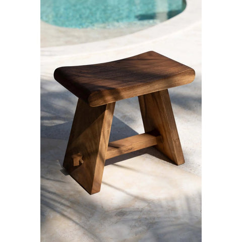 The Suar Stool - 50, with its curved seat and angled legs, sits on a sunlit patio near the edge of a swimming pool.