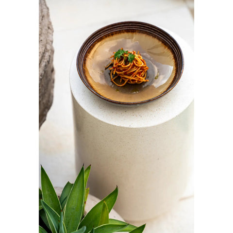 A set of four Comporta Pasta Plates (large), crafted from Portuguese ceramics, features brown rims and displays spaghetti with tomato sauce and herbs, resting on a round white pedestal beside a green leafy plant.