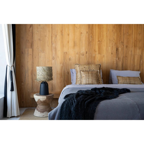 A modern bedroom with a wooden accent wall, gray bedding, woven pillows, and The Kona Stool - Natural as a side table beside the bed. A woven lampshade sits nearby while light streams in through white curtains on the left.