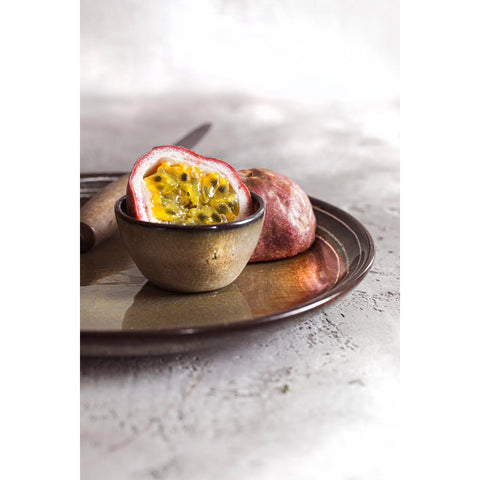 A halved passion fruit with bright yellow pulp and black seeds sits in a small brown bowl of handmade tableware on a round plate; a whole passion fruit and a knife are in the background on a light surface.