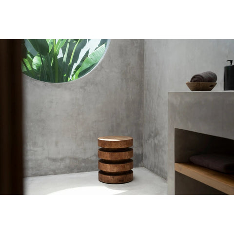 A minimalist bathroom with concrete walls, a round window framing greenery, and a stack of The Massaka Stool - Natural on the floor next to a built-in shelf holding rolled towels and toiletries.