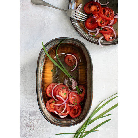 A rustic bowl with a crackle glaze finish holds sliced tomatoes and red onions, garnished with green herbs. Nearby, another bowl—crafted from Portuguese ceramics—contains the same salad and a fork, all set on a light surface with herb sprigs.