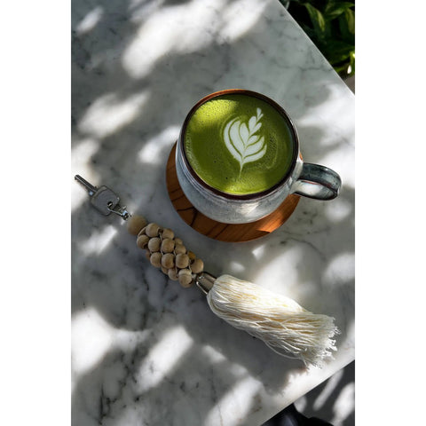 A cup of matcha latte with leaf latte art sits on a marble table, served in handmade tableware, beside a decorative keychain with wooden beads and a long white tassel. Sunlight and shadows fall across the scene.