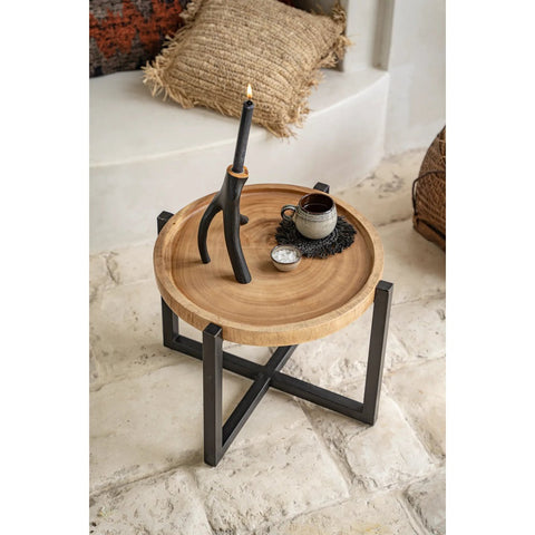 A round wooden side table with black metal legs holds a black candle in a unique holder, a black cup and saucer of handmade tableware, and a small round object. A textured pillow and woven basket are in the background on a stone floor.