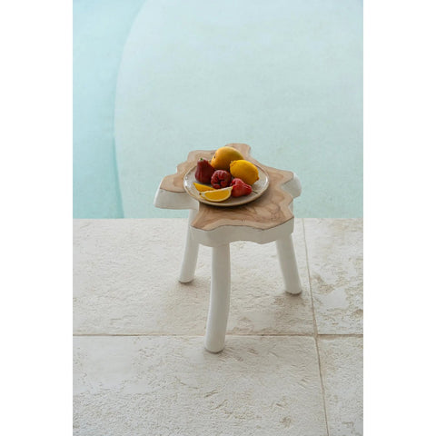 A small, white three-legged wooden stool holds a plate of lemons and strawberries made from handmade tableware, positioned on light stone tiles next to a light blue swimming pool.