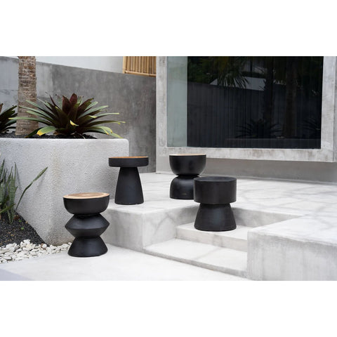 Four modern charcoal stools with wooden tops are arranged on concrete steps and a patio in a minimalist outdoor setting, featuring potted plants and a large window in the background.