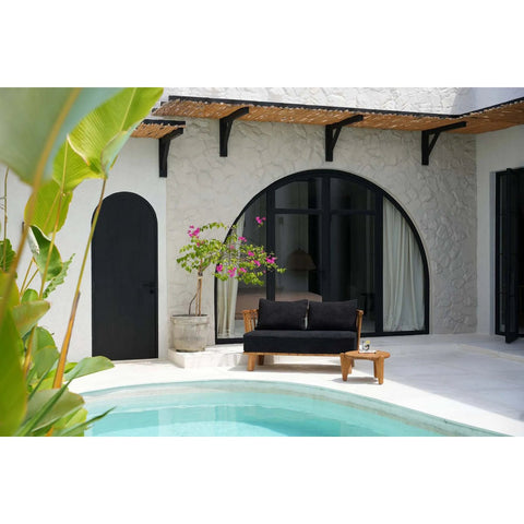 A modern patio features a small pool, black-cushioned wooden sofa, The Seseh Coffee Table - S - Outdoor, potted plants, and white textured walls with large arched windows and a door. Sunlight fills the tranquil outdoor space.