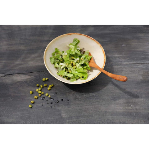 A ceramic bowl of mixed green salad with a wooden spoon rests on a dark wooden surface, showcasing elegant handmade tableware with scattered green peas and droplets nearby.
