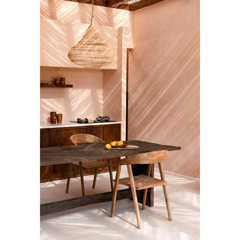 A sunlit, minimalist kitchen features wooden furniture, a dark wooden table, and an elegant dining chair paired with a woven pendant lamp. Lemons rest on the table as light creates striped patterns across the walls and surfaces.