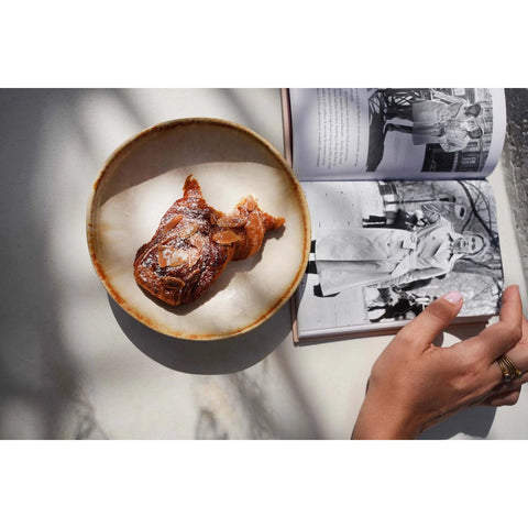 A hand holds open a book with black-and-white photos beside The Cascais Appetizer Plate - Set of 6, showcasing a fish-shaped pastry dusted with powdered sugar on a white surface as soft shadows fall across the scene.