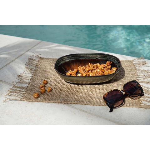 A bowl of caramel popcorn in Portuguese ceramics with a crackle glaze finish sits on a woven placemat by the swimming pool, sunglasses nearby. Sunlight casts shadows on the placemat and white poolside tiles.