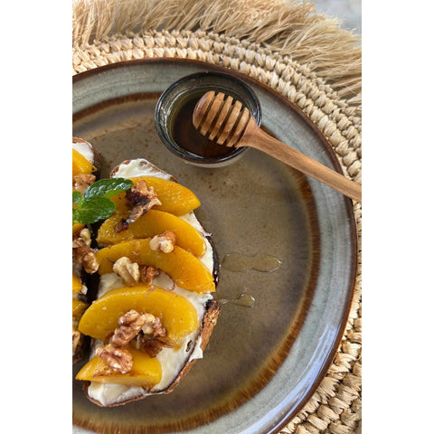 A plate with bread topped with cream, peach slices, walnuts, and mint sits on a woven placemat. The scene is completed by a small bowl of honey with a honey dipper, served on elegant handmade tableware inspired by Portuguese ceramics.