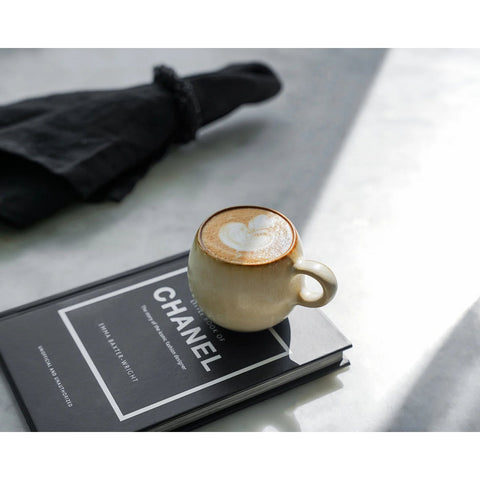 A cappuccino with heart-shaped latte art sits on a Chanel book, served in The Cascais Espresso Cup - S (Set of 6), paired with chip-proof Portuguese ceramics and a folded black napkin and utensils on a light marble surface.