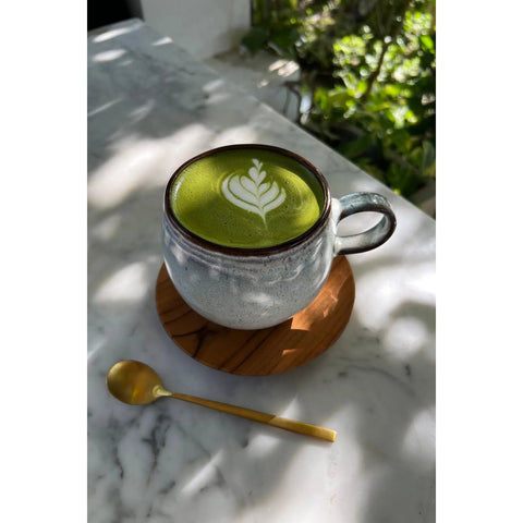 A ceramic cup of matcha latte with leaf-shaped latte art sits on a wooden coaster next to a gold spoon, atop a marble table. Sunlight filters in, illuminating the handmade tableware and lush green plants in the background.