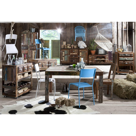 A rustic room with wooden walls and floors, mismatched colorful chairs, a wooden dining table, the Industrial Low Board Woodz, vintage cabinets, a hammock, hay bales, and sunlight streaming through an open window.