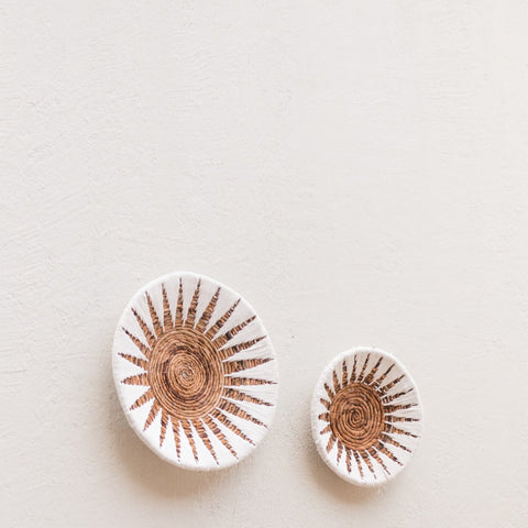 Two round Decorative Bowl MADALA baskets made of woven banana fibre with sunburst patterns hang asymmetrically on an off-white textured wall. Their white and brown details add a unique boho charm, available in two sizes.