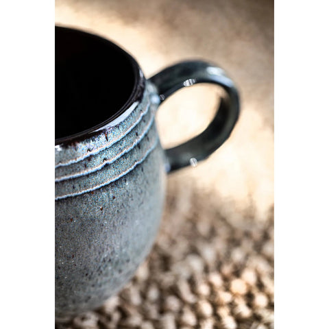 Close-up of a textured ceramic mug with a handle, featuring a blue-gray crackle glaze. The background is softly blurred, highlighting the mug’s surface details and matte finish—a beautiful example of handmade tableware.