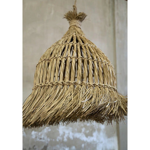 A close-up of The Guagua Pendant - Natural - L, a handmade grass lamp with a conical woven straw shade and fringed edges, hanging against a textured gray wall.