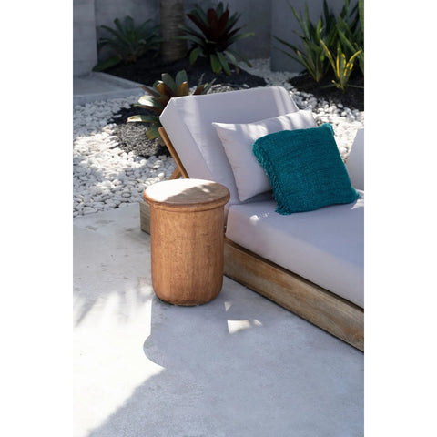 The Killa Stool - Natural sits beside a cushioned outdoor lounge chair with a teal pillow, arranged on a concrete patio bordered by white pebbles and lush green plants.