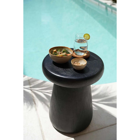 A round black side table by a pool holds a glass of water with a lime slice, a bowl of salad, and two small bowls with snacks served in elegant Portuguese ceramics. Sunlight casts shadows on the floor beside the table.