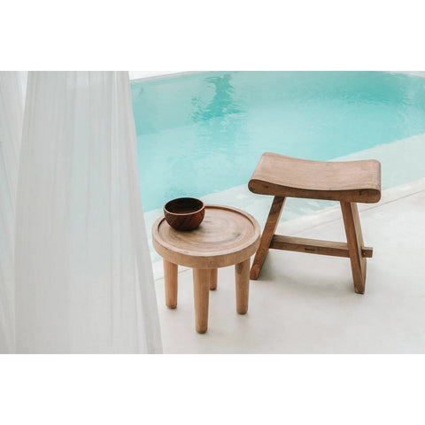 A small round wooden table with a bowl sits next to The Suar Stool - 50 by a clear blue pool, with sheer white curtains partly visible on the left.