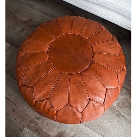 The Leather Pouffe - Cognac Shiny XL features a round, handcrafted design with visible petal-like stitching and sits on a wooden floor beside the edge of a white bed.