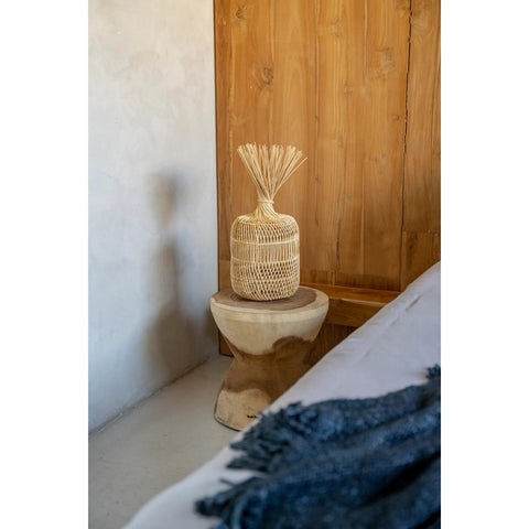 A woven basket with straw accents rests on The Kona Stool - Natural, crafted from Indonesian suar wood, in a cozy room with wooden panel walls and a light blanket draped over the bed.