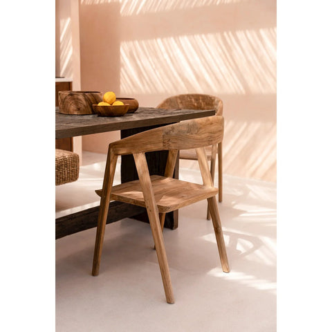 An elegant dining chair is placed next to a dark wooden table with a bowl of lemons on top. Sunlight streams through slats, casting patterned shadows on the pale floor and wall.