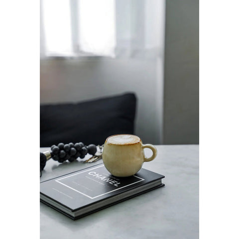 A set of six beige Cascais Espresso Cups, made from chip-proof Portuguese ceramics, rests on a black and white Chanel book beside a beaded accessory on a light gray table with a blurred pillow and window in the background.