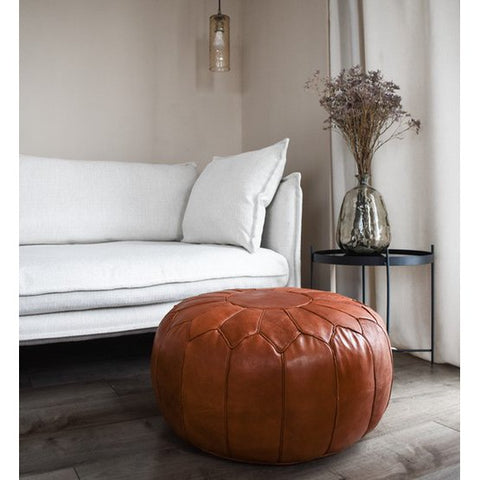 A cozy living room with a white sofa, a Leather Pouffe - Cognac Shiny XL on wooden flooring, a small black side table with a glass vase of dried flowers, and a hanging pendant light.