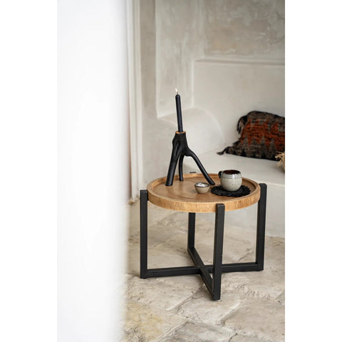 A round wooden side table with black metal legs holds a black candle in a sculptural holder, a small bowl of handmade tableware, and a cup. A pillow rests on a built-in seat in the beige, minimalist room.