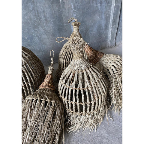 The Guagua Pendant - Natural - L features a handmade, dome-shaped woven design with fringed edges and looped handles, resembling a grass lamp. Displayed on concrete against a gray textured wall for an artisanal touch.