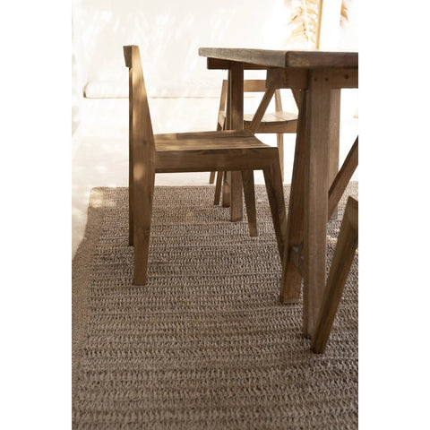 The Paxi Dining Chair - Natural - Outdoor sits on a woven beige rug in a sunlit room with soft, natural light and minimal decor.