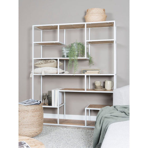 A modern white metal and wood shelving unit stands against a gray wall, adorned with books, plants, baskets, blankets, and vases. In the foreground are a sofa and the Poof Anne decorative ottoman.