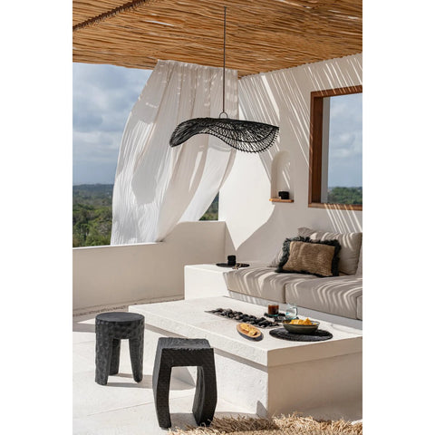 A modern outdoor patio with a white sofa, neutral pillows, sheer white curtains, two black stools, a coffee table with snacks and drinks, and The Chapeau Pendant - Black - L as the statement light, all overlooking greenery beneath a partly cloudy sky.