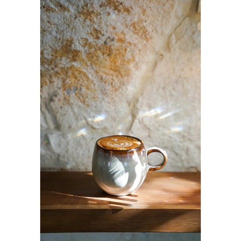 A ceramic mug with latte art sits on a wooden surface, illuminated by soft sunlight against a textured stone wall in the background—an inviting piece of handmade tableware.