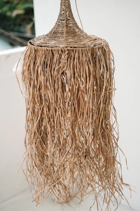 A handcrafted tropical destination lamp made of woven natural fibers hangs from the ceiling. It has a conical top and long, frayed, tassel-like strands cascading downward, creating a rustic, bohemian look. The background is softly blurred.