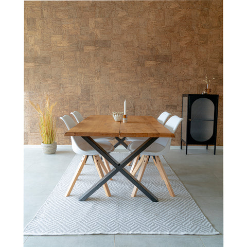 A modern dining room with a wooden table and four white chairs. The table has metal X-shaped legs and is placed on a patterned rug. A small black cabinet stands against a textured wall, and a vase with dried grass is beside it.