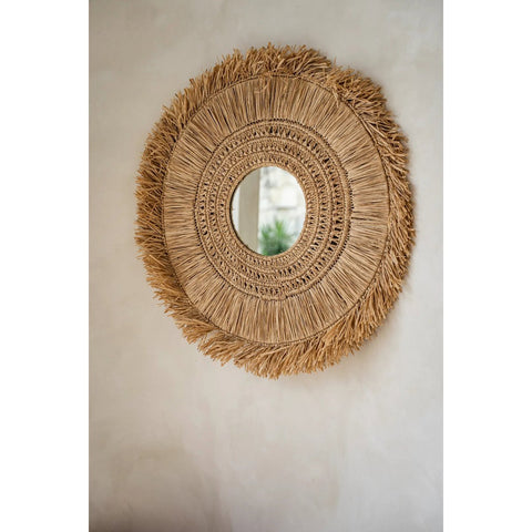 Round woven mirror with fringe details on a plain wall