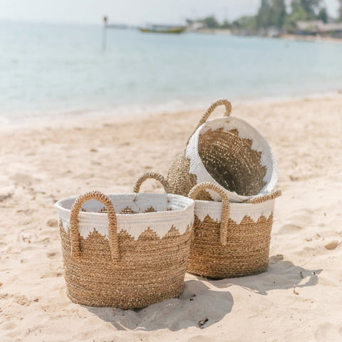 Three Round Seagrass Baskets WAIGEO with white painted tops are placed on a sandy beach near the shore, with the sea and distant trees in the background.