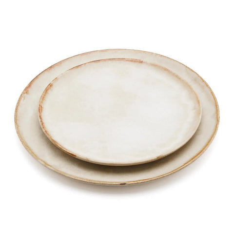 Two round, ceramic plates with a rustic, off-white finish and slightly uneven, brownish edges showcase handmade tableware inspired by Portuguese ceramics, stacked with the smaller plate on top of the larger one.