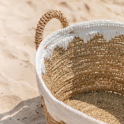 A close-up of the Round Seagrass Baskets WAIGEO, featuring a handmade design with a white decorative fringe along the rim, resting on sandy ground with sunlight creating soft shadows.