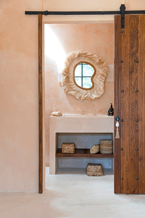 A bathroom with a rustic, boho style. It features a wooden sliding door, a mirror with a fringed, circular design, and a beige countertop. Below are woven baskets on a shelf, with a small towel and toiletries on the counter.