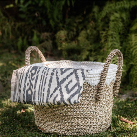 A Round Seagrass Basket WAIGEO with rope handles sits on grass, holding a white and black patterned cloth draped over its edge, with greenery and ferns softly blurred in the background.